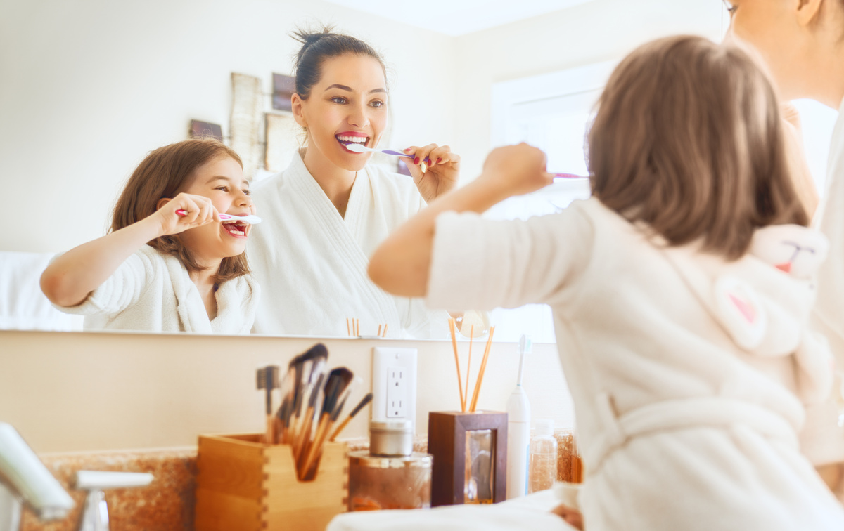 Mulher e criança escovando os dentes juntas, em frente ao espelho. 