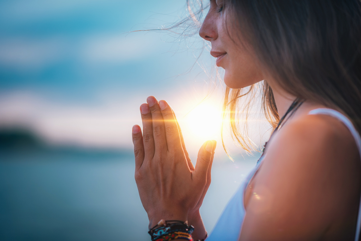 Mulher com as mãos juntas em frente ao corpo enquanto a luz do sol a atravessa, mostrando meditar, prática que ajuda a alcançar o equilíbrio do chakra coronário.