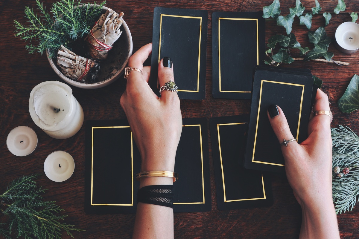 Mãos enfileirando cartas de Tarot de design preto em cima de mesa de madeira escura, ao lado de ervas e velas.