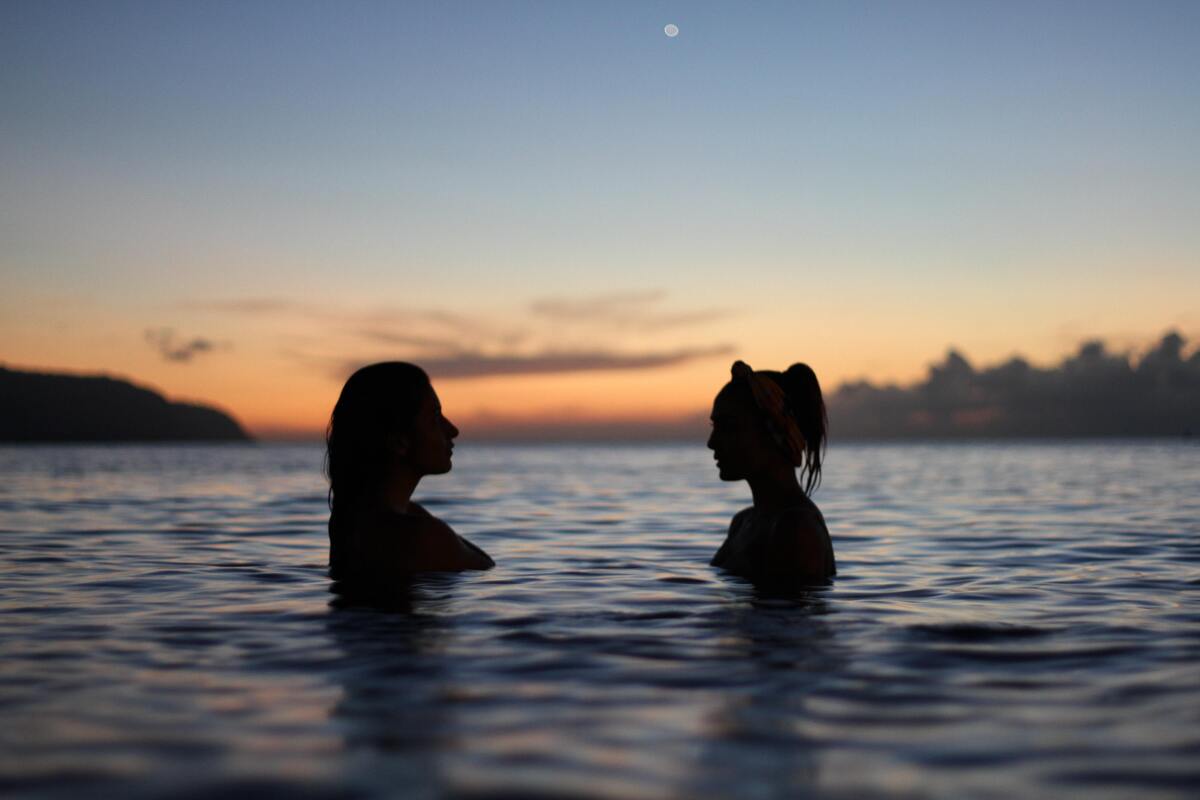 Silhueta de duas mulheres na água, montanhas ao fundo. 