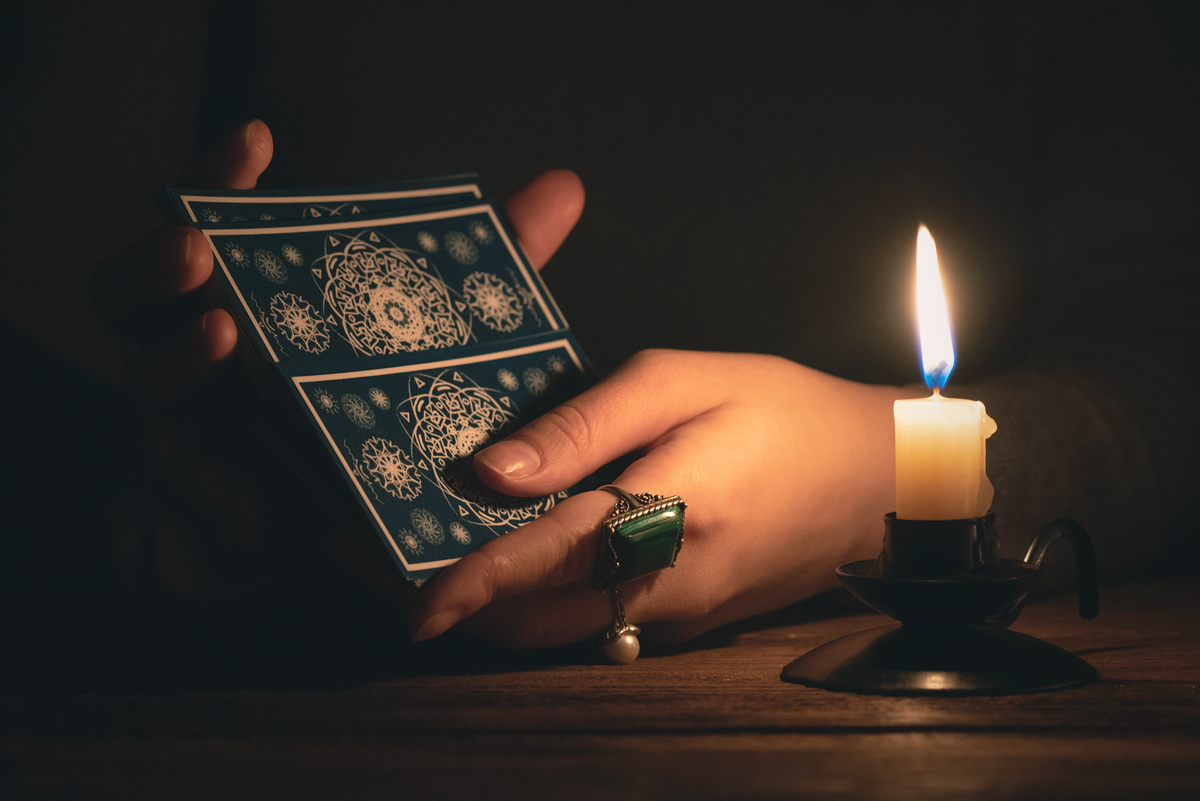 Mãos com anéis embaralhando cartas de Tarot em cenário escuro, iluminado apenas por uma vela.