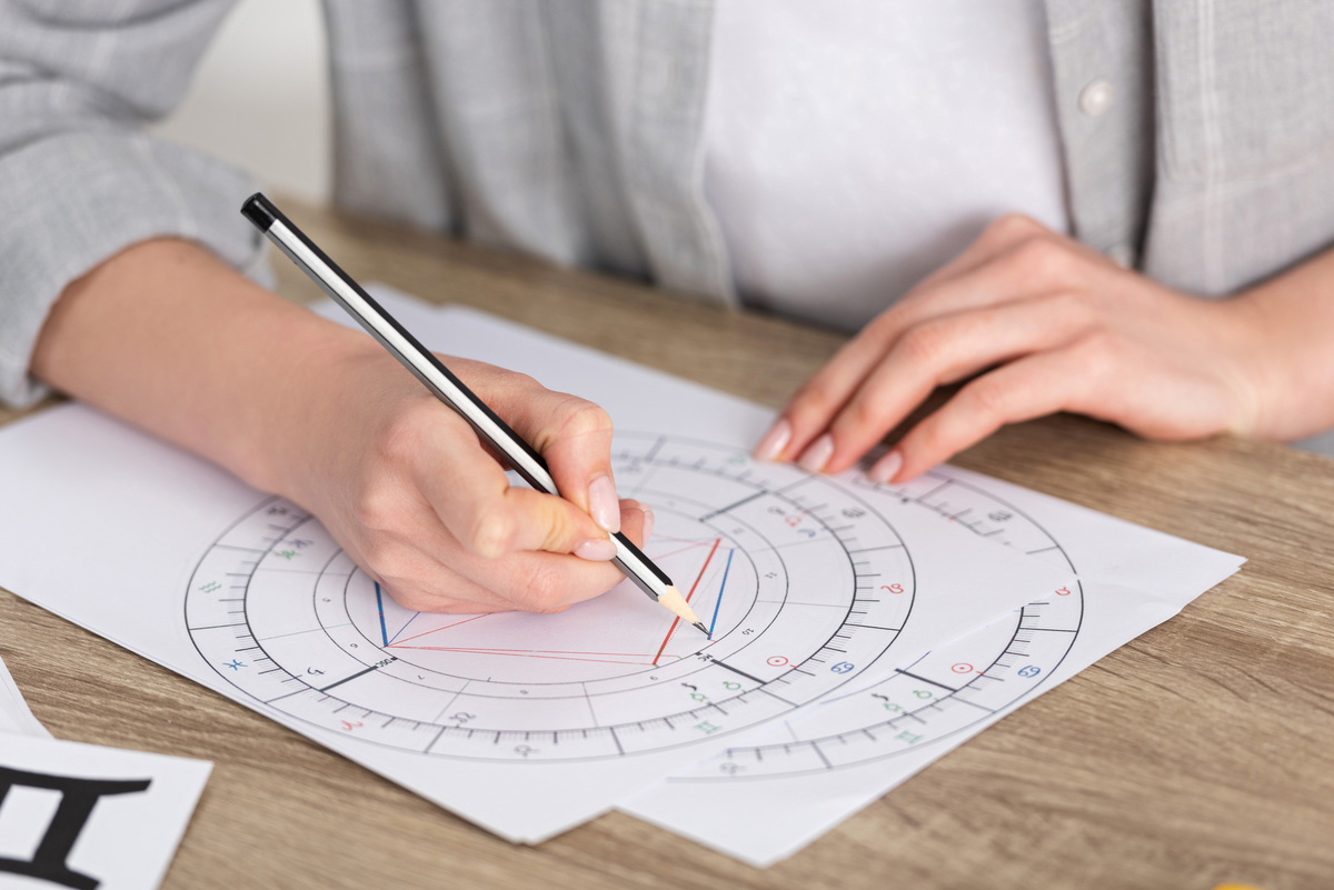 Pessoa utilizando um lápis para desenhar as angulações em uma folha com o mapa astral impresso, indicando o estudo das casas e posicionamento dos astros.
