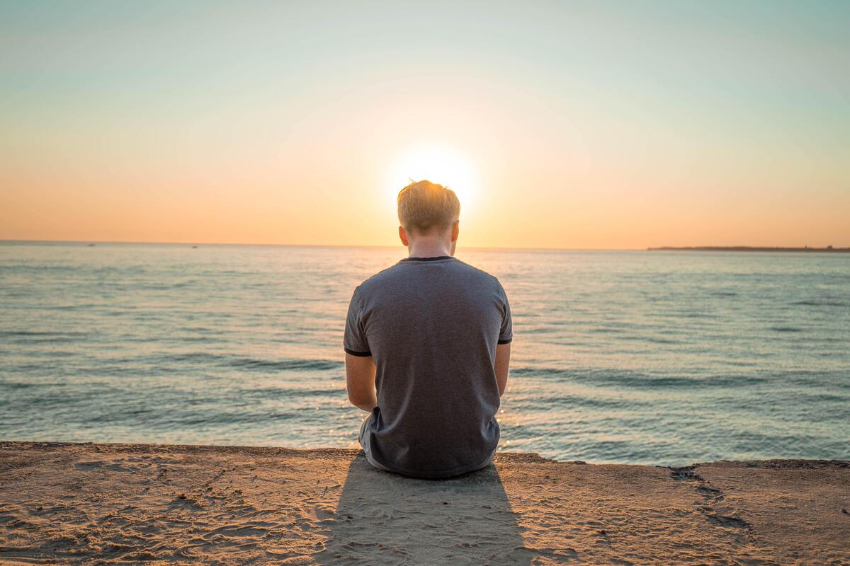 Homem sentado na praia, de costas, com o horizonte ao fundo. 