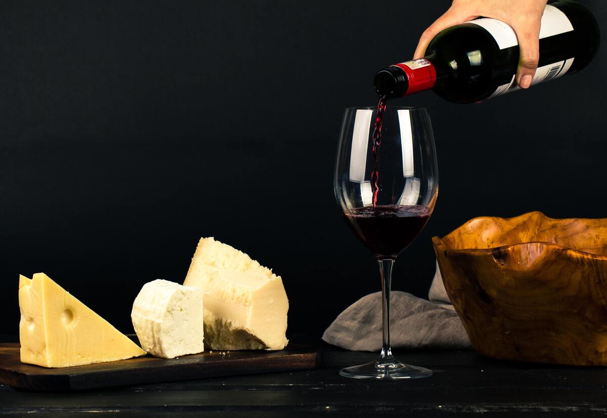 Mão com garrafa de vinho derramando dentro de uma taça e queijos à sua volta