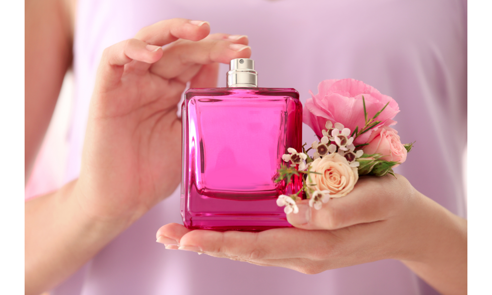 Mulher segurando um fraco de perfume rosa nas mãos e algumas flores.