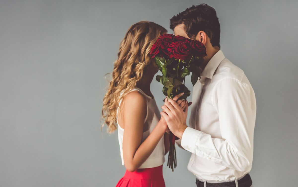 Casal de homem e mulher se beijando atrás de buquê de rosas.