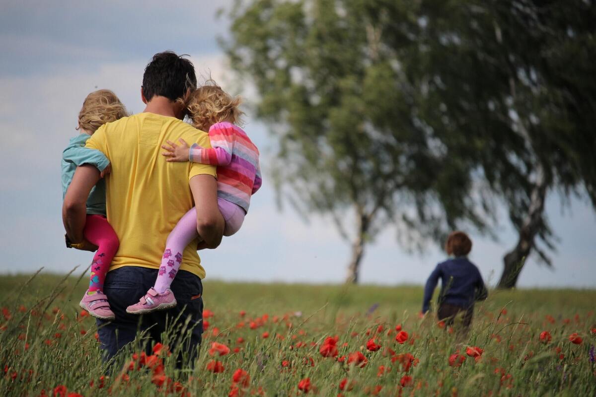 Homem carregando duas crianças em um campo gramado.