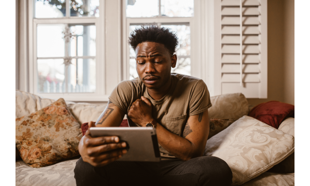 Homem negro chorando sentado em um sofá com um tablet e uma mão apoiada em seu peito.