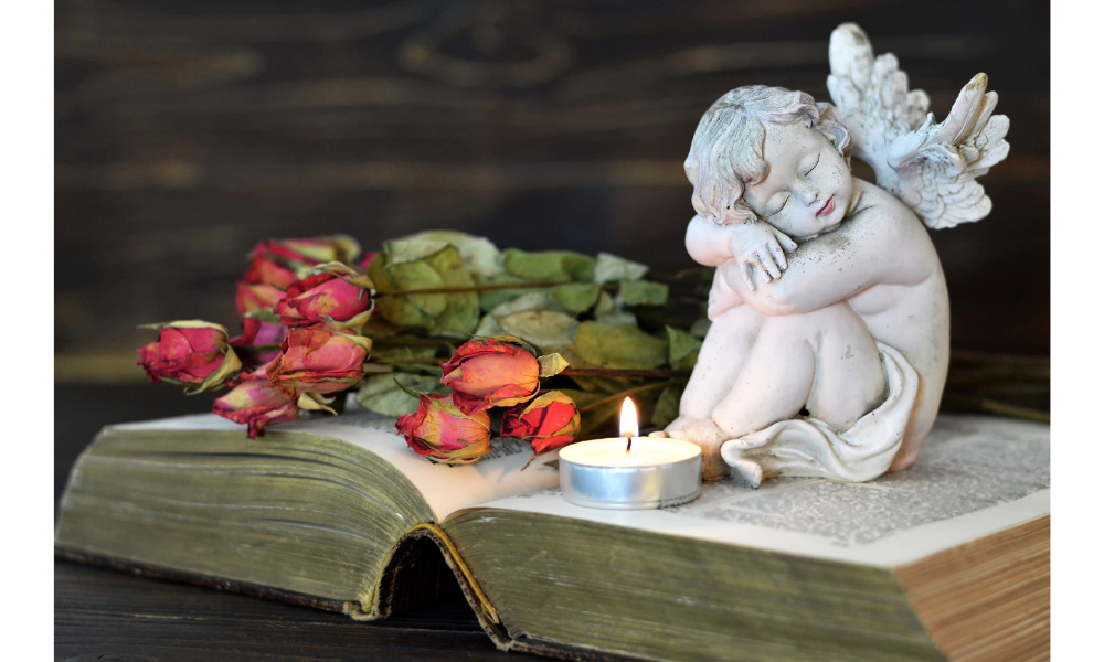 Imagem de um anjo sobre uma bíblia, junto de flores secas e vela.