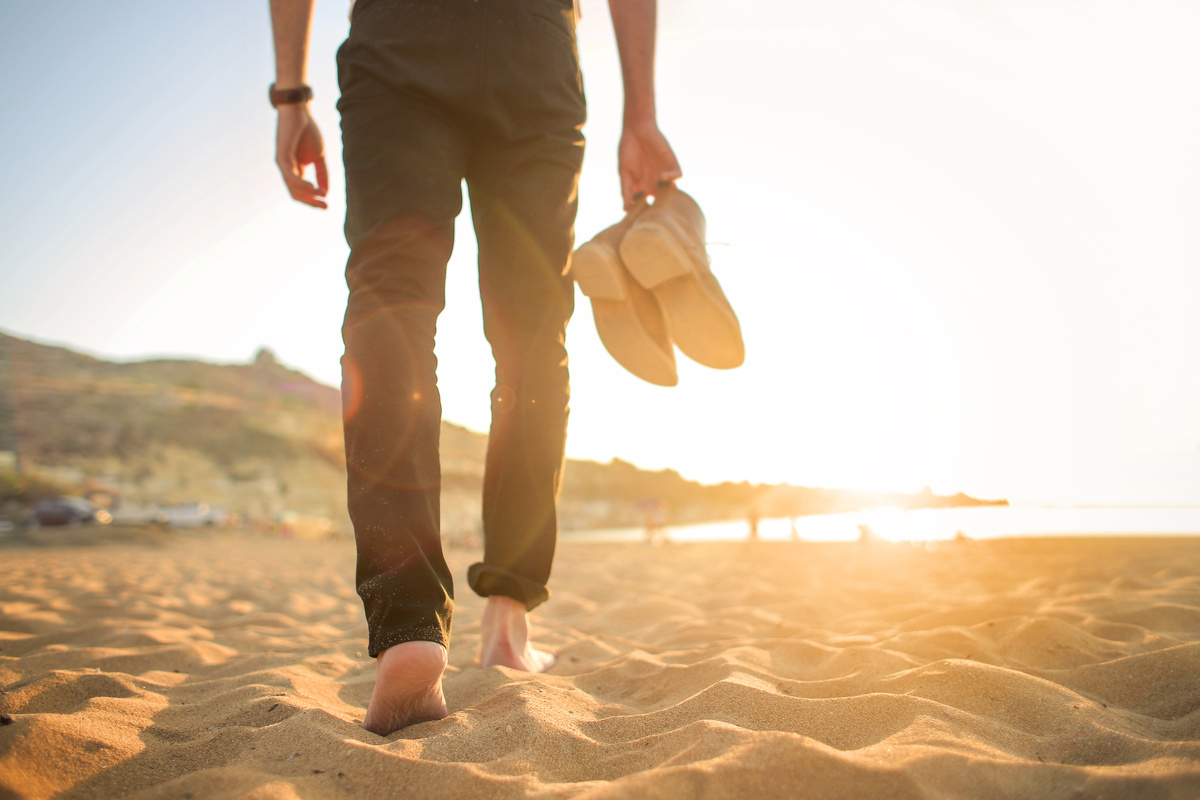 Homem caminhando descalço na praia, enquanto segura seus sapatos.