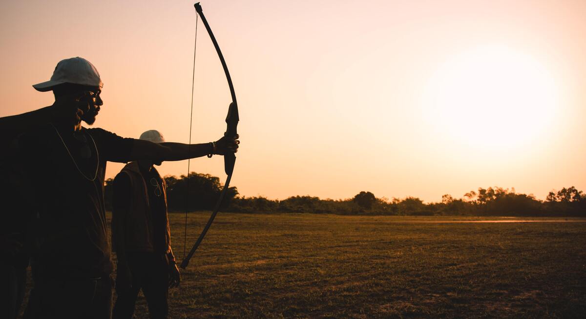 Silhueta de dois homens com arco e flecha em um campo gramado.