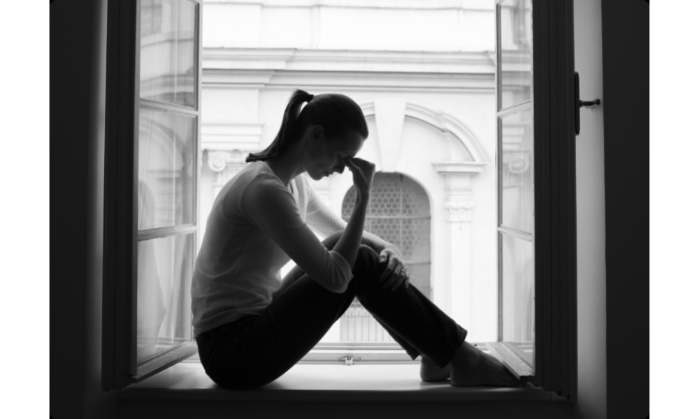 Imagem preta e branca com mulher triste sentada em uma janela com uma mão na testa.