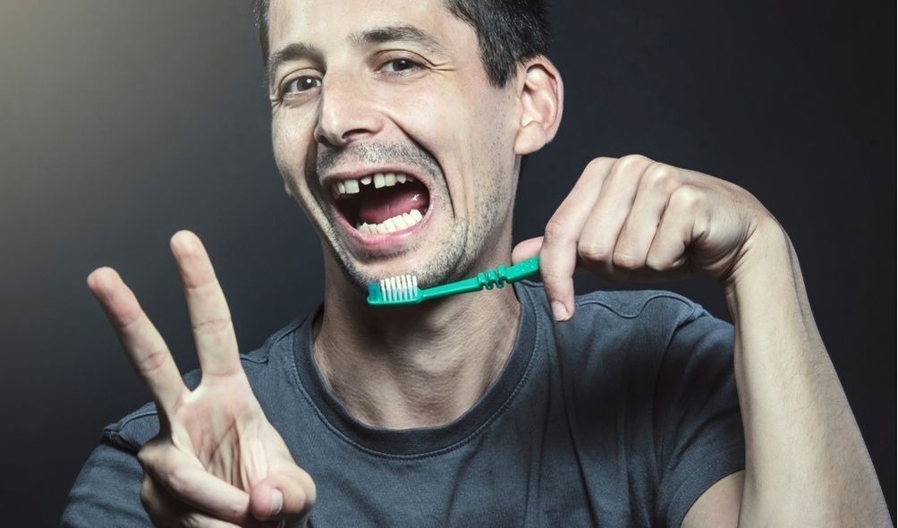 Homem com dentes faltando, fazendo um sinal de paz, e segurando uma escova de dentes