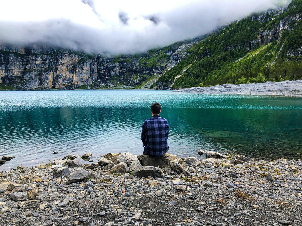 Homem de costas, sentado na beira de um lago. 