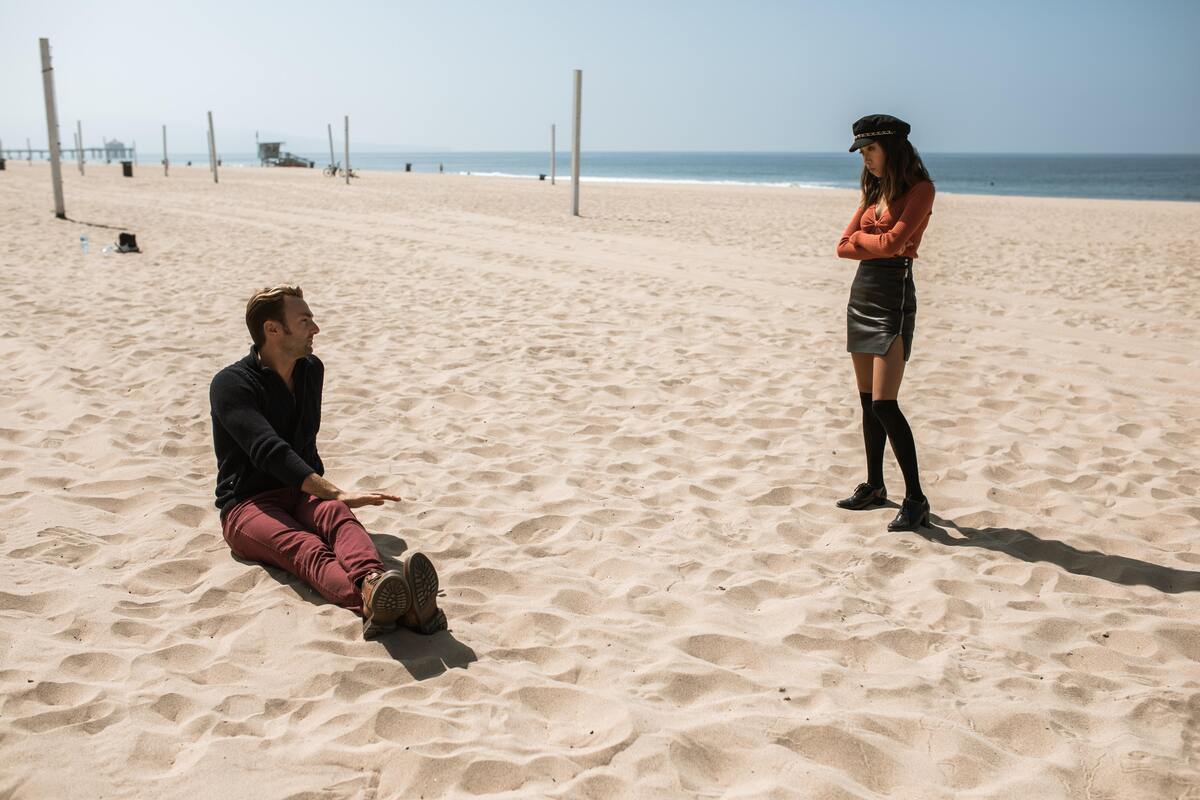 Um homem sentado na areia e uma mulher em pé na areia