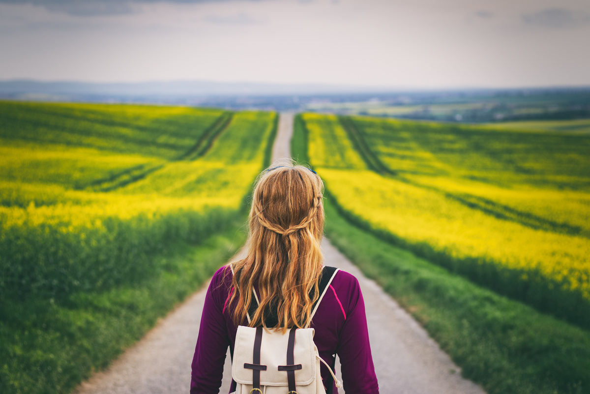 Mulher focada no seu caminho, andando em uma estrada de terra entre dois campos verdes.