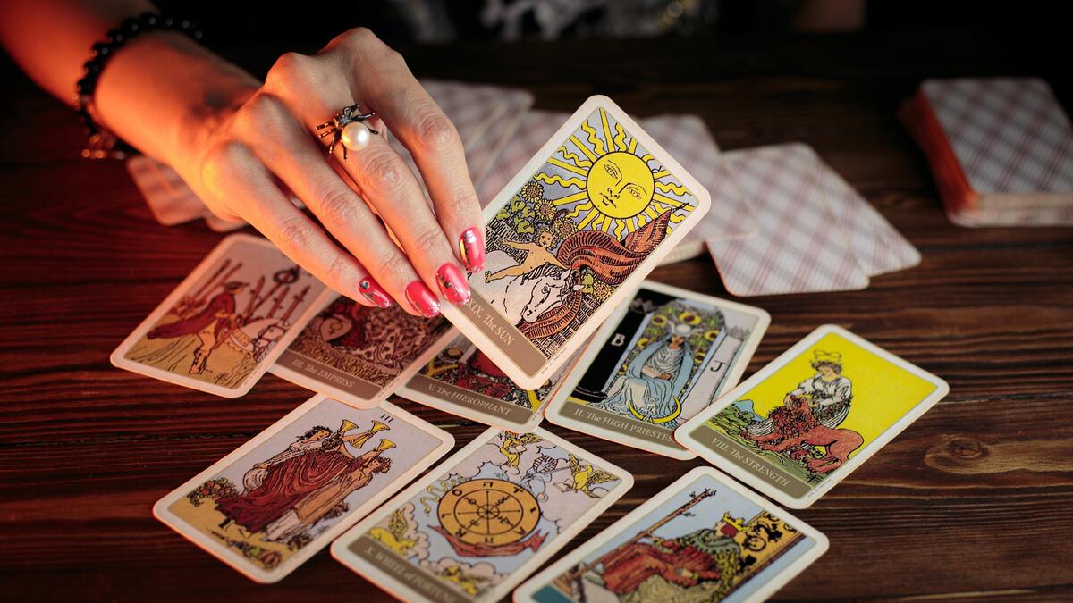 Mão feminina pegando uma carta de Tarot, outras cartas jogadas na mesa. 