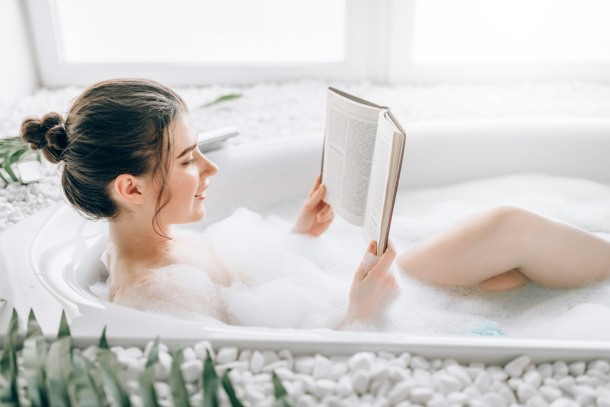 mulher tomando banho de banheira e lendo livro.
