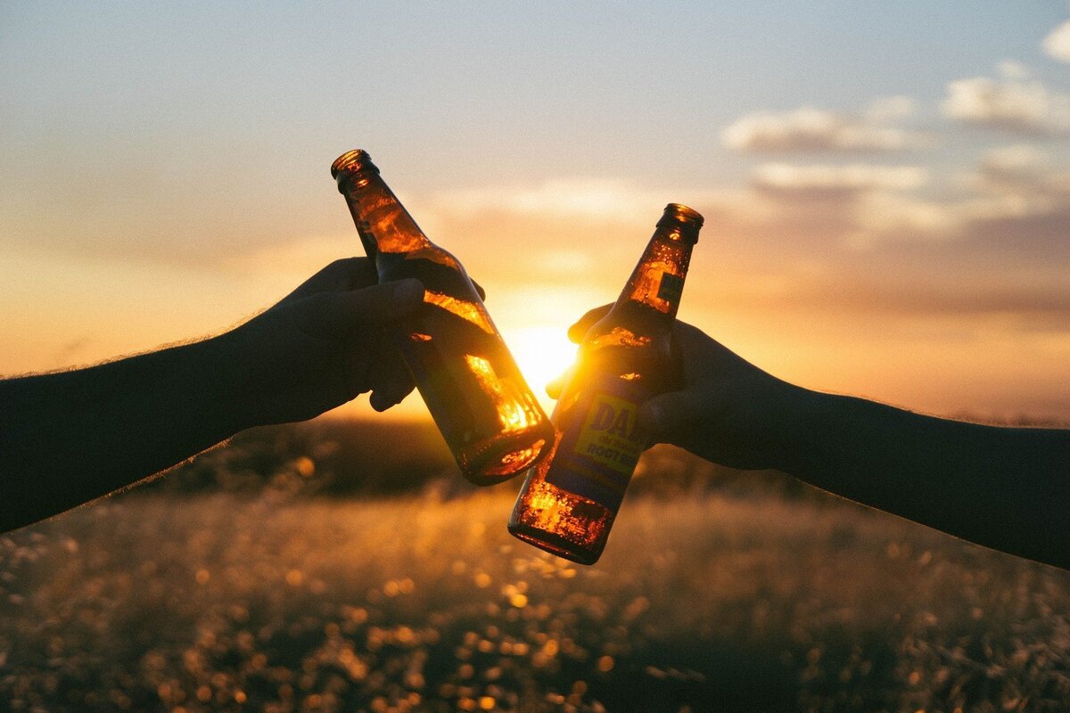 Duas pessoas brindando com garrafas no pôr do sol