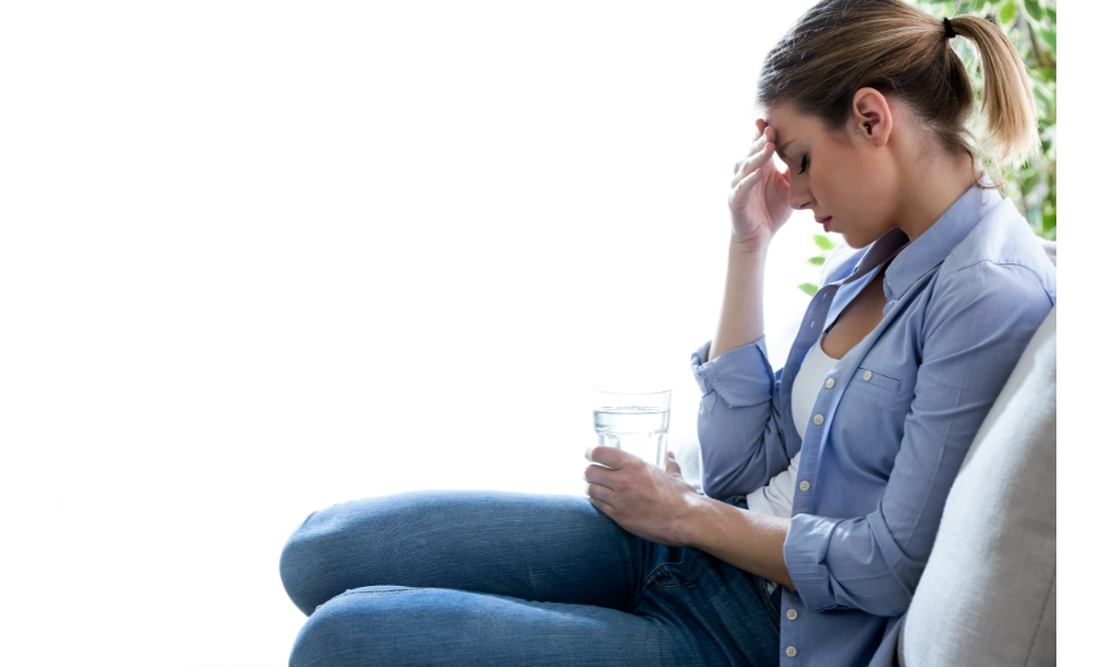 Mulher sentada com dor de cabeça e segurando um copo de água.