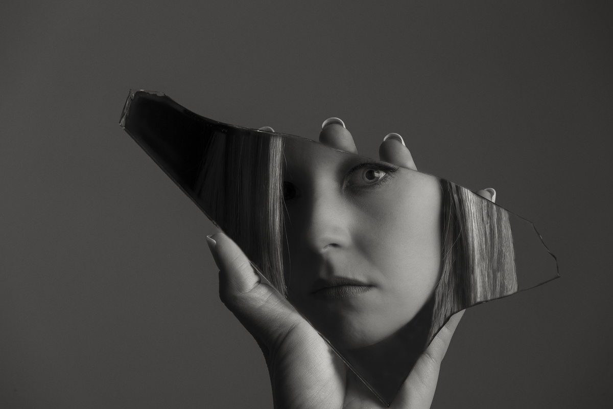 Imagem em preto e branco de uma mulher olhando o seu reflexo em espelho quebrado.